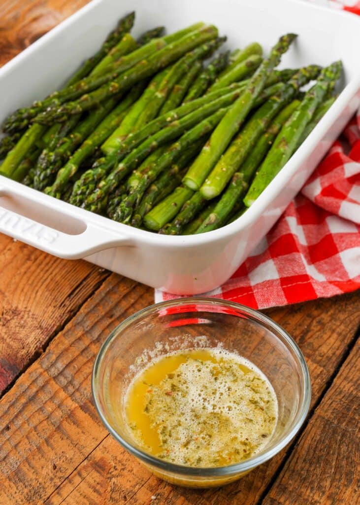 a white baking dish is loaded with roasted asparagus spears. beside it on a wooden tabletop there is a small glass bowl with melted garlic butter sauce.
