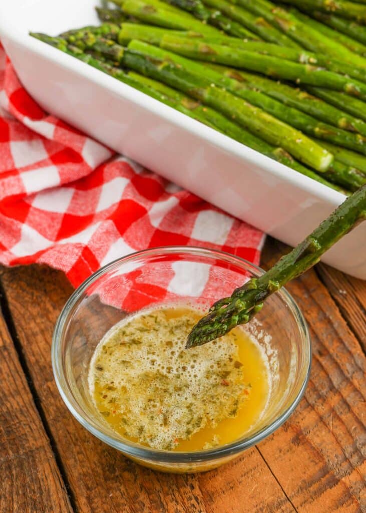 A clear glass bowl with melted compound butter in it and a spear of asparagus is being dipped in the butter.