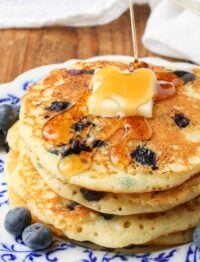A close up shot of drizzling maple syrup over a pat of butter atop a pile of buttermilk blueberry pancakes