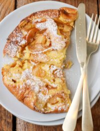 a top down photo of a white plate with part of a dutch baby with sliced apples and powdered sugar, a knife and fork resting on the plate beside the food