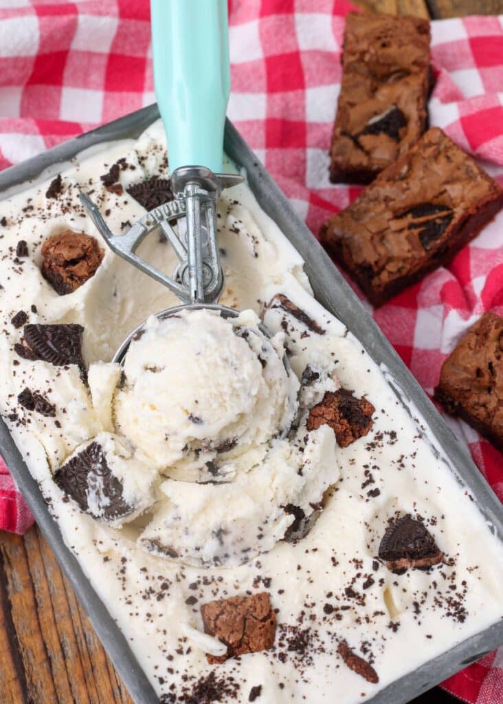 a scoop with a cyan handle is stuck into a freezer safe container full of oreo brownie ice cream with brownies visible in the background
