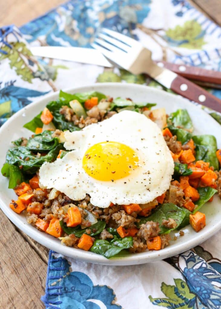 Sausage sweet potato hash with a sunny side up egg, served in a white bowl with a blue and white floral pattern towel