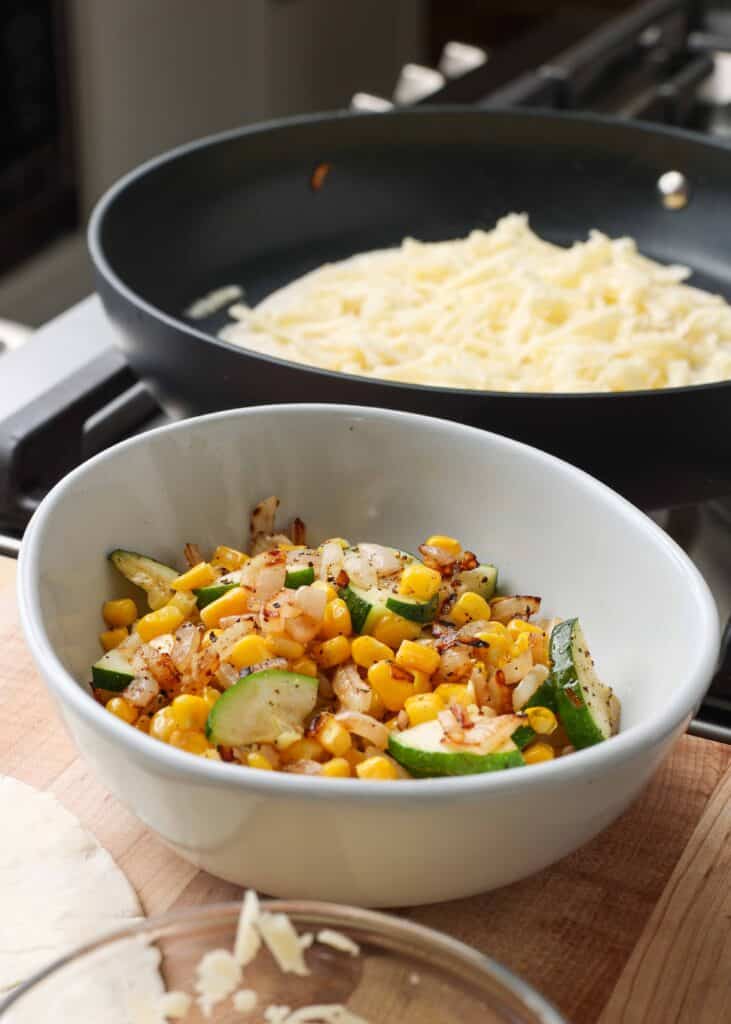 Shredded white cheese piled in a black bowl and diced zucchini and corn tossed in a white bowl