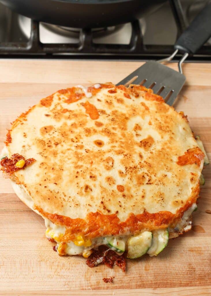 Overhead shot of double quesadilla with golden flour tortillas, green zucchini, cheese, and corn, served on a light brown wooden cutting board with a metal spatula